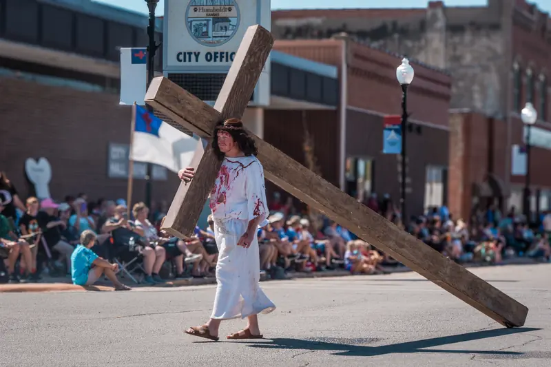A man portraying Jesus carries a heavy wooden cross along the parade route as spectators watch.