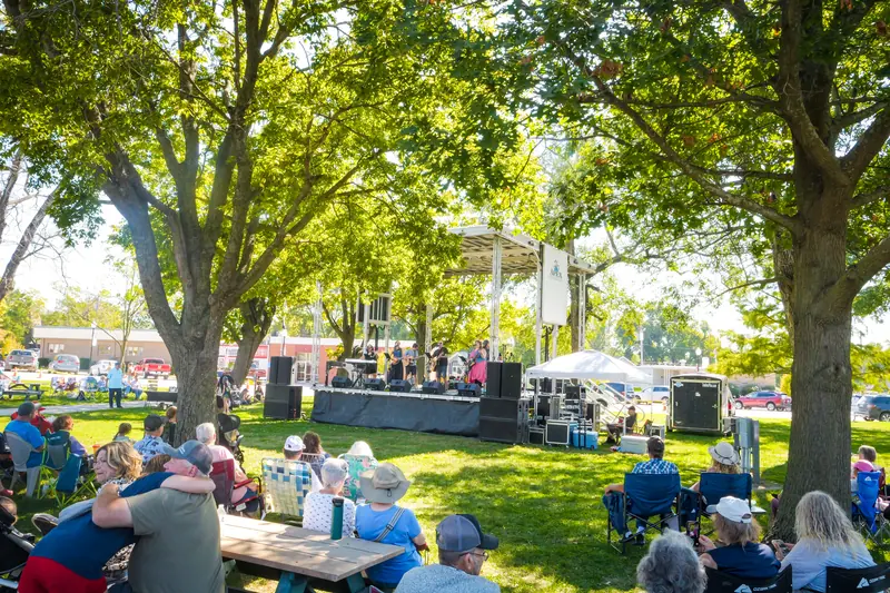Listeners in lawn chairs under shade trees face the music stage on the Town Square.